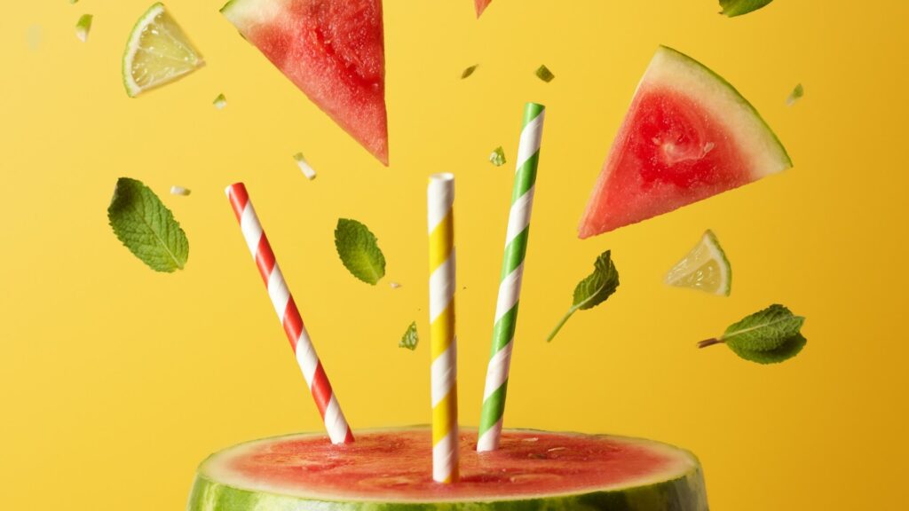 Watermelon slices, mint leaves, and lime wedges floating around colorful straws in a carved watermelon, capturing a playful summer cocktail vibe.