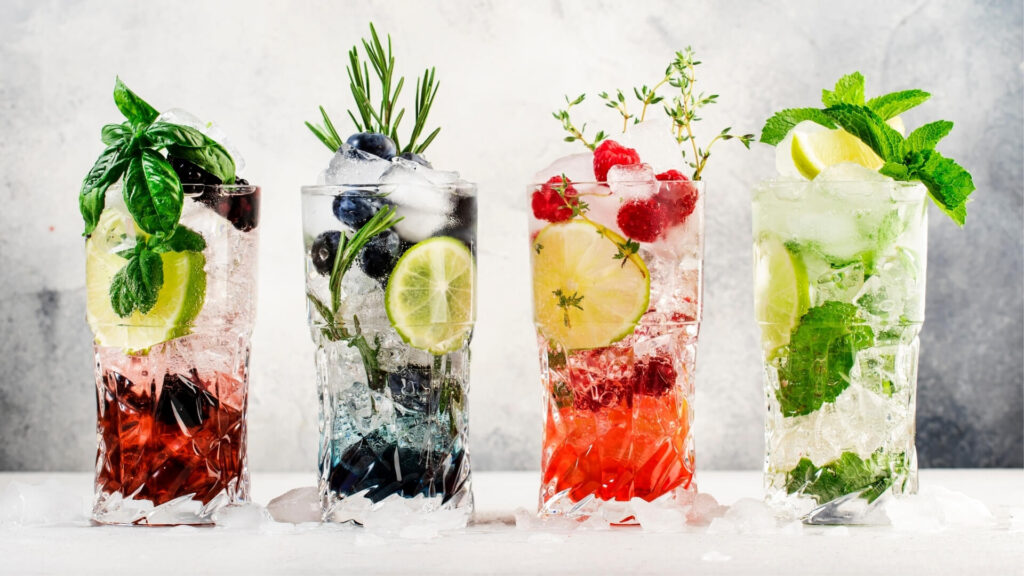 Colorful infused cocktails in highball glasses garnished with herbs, lime, and fresh berries on ice.