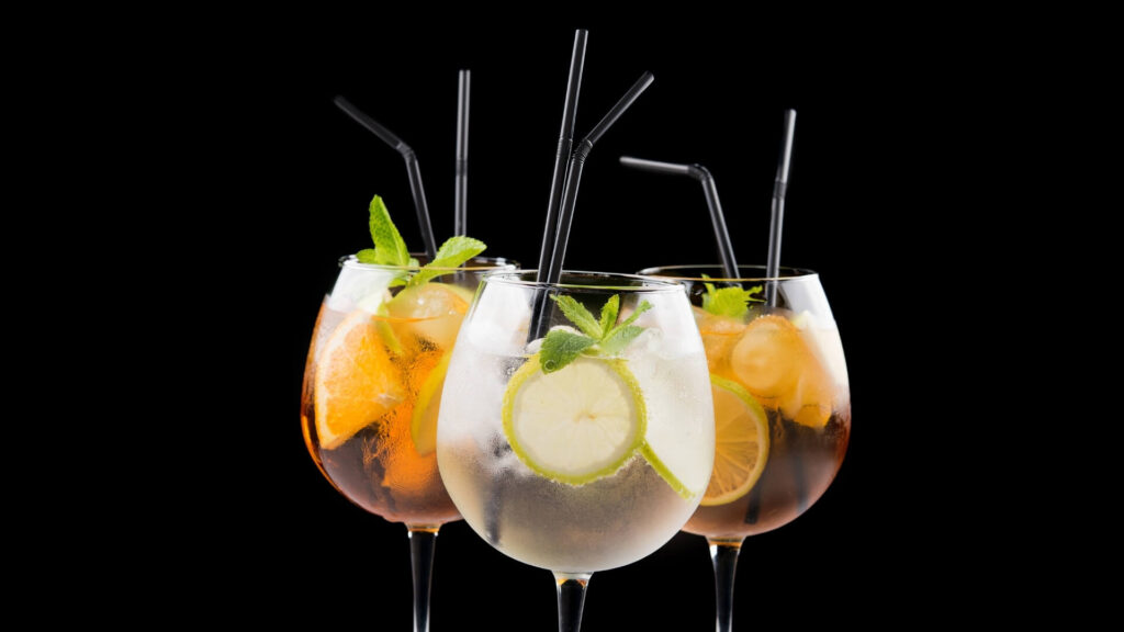 Three stemmed wine glasses filled with Spritz cocktails, lime slices, mint leaves, and black straws, set against a black background.