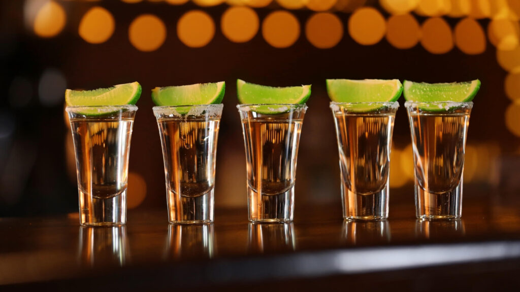 Five tequila shot glasses with lime wedges and salted rims lined up on a bar counter